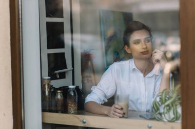 Güzel, genç bir kız, elinde bir fincan kahveyle bir kafede oturur ve pencereden dikkatlice dışarı bakar. Sokak yansımalı cam içinden fotoğraf. Yumuşak seçici odak.