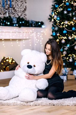 Yeni yıl ağacının yanında genç ve güzel bir kız büyük beyaz bir oyuncak ayıyla neşeyle gülümsüyor. Mutlu Noeller ve tatiller. Yumuşak seçici odak.