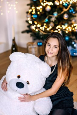 Yeni yıl ağacının yanında genç ve güzel bir kız büyük beyaz bir oyuncak ayıyla neşeyle gülümsüyor. Mutlu Noeller ve tatiller. Yumuşak seçici odak.