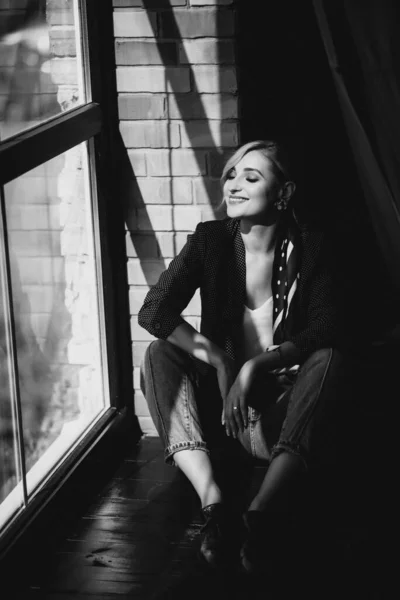 Beautiful young woman blonde in jeans with a polka dot jacket and a scarf in her hair in a loft studio near the window with sunbeams. Soft selective focus. Black and white photo.