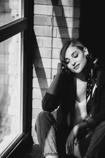 Beautiful young woman blonde in jeans with a polka dot jacket and a scarf in her hair in a loft studio near the window with sunbeams. Soft selective focus. Black and white photo.