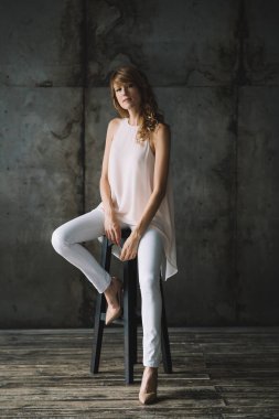 Beyaz pantolonlu, kıvırcık saçlı, güzel, genç, narin, sarışın bir kadın bir stüdyo dairede bir bluz ve ayakkabıyla bir bar taburesinde oturuyor. Yumuşak seçici odak.