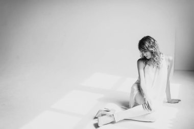 Beyaz pantolonlu, kıvırcık saçlı, güzel, genç, ince, sarışın bir kadın, stüdyoda bir bluz ve ayakkabılar beyaz bir arka planda oturuyor. Yumuşak seçici odaklanma. Siyah beyaz fotoğraf.