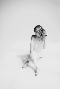 Beyaz pantolonlu, kıvırcık saçlı, güzel, genç, ince, sarışın bir kadın, stüdyoda bir bluz ve ayakkabılar beyaz bir arka planda oturuyor. Yumuşak seçici odaklanma. Siyah beyaz fotoğraf.