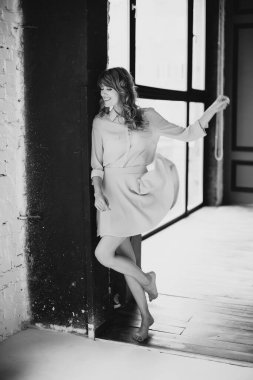 Güzel, narin, sarışın, kıvırcık saçlı bir kadın. Pencere kenarında dans eden bir stüdyo dairede çıplak ayaklı, gülümseyen bir kadın. Yumuşak seçici odaklanma. Siyah beyaz fotoğraf.