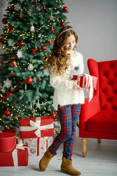 長いブロンドの髪をした小さな美しい女の子は 新年のインテリアのクリスマスツリーの横に立っており 彼女の手に贈り物と赤い箱を保持しています — ストック写真