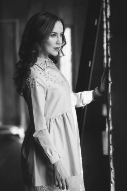 Kıvırcık siyah saçlı ve pudralı elbiseli kırmızı dudaklı güzel bir genç kadın çatı katındaki stüdyonun penceresinin yanında duruyor. Yumuşak seçici odaklanma. Siyah beyaz sanat fotoğrafı..