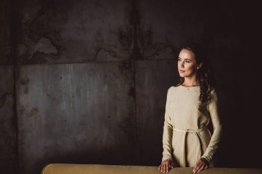 Koyu kıvırcık saçlı, kahverengi gözlü, güzel, genç, esmer bir kadın çatı katında altın bir elbise içinde uzaklığa bakıyor. Yumuşak seçici odak.