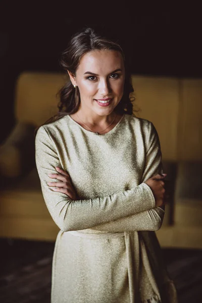 Koyu renk kıvırcık saçlı, kahverengi gözlü, altın rengi elbisesi olan ve tavan arasındaki hardal kanepenin arka planında duran güzel, genç, esmer bir kadın kameraya neşeli bir şekilde bakıyor. Yumuşak seçici odak.