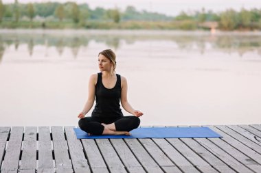 36 yaşında beyaz bir kadın, sabahın köründe, nehrin kenarındaki ahşap bir rıhtımda lotus pozisyonunda yoga yapıyor. Sağlıklı yaşam tarzı kavramı. Sabah meditasyonu, rahatlama.