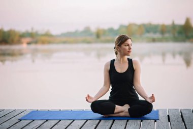 36 yaşında beyaz bir kadın, sabahın köründe, nehrin kenarındaki ahşap bir rıhtımda lotus pozisyonunda yoga yapıyor. Sağlıklı yaşam tarzı kavramı. Sabah meditasyonu, rahatlama.