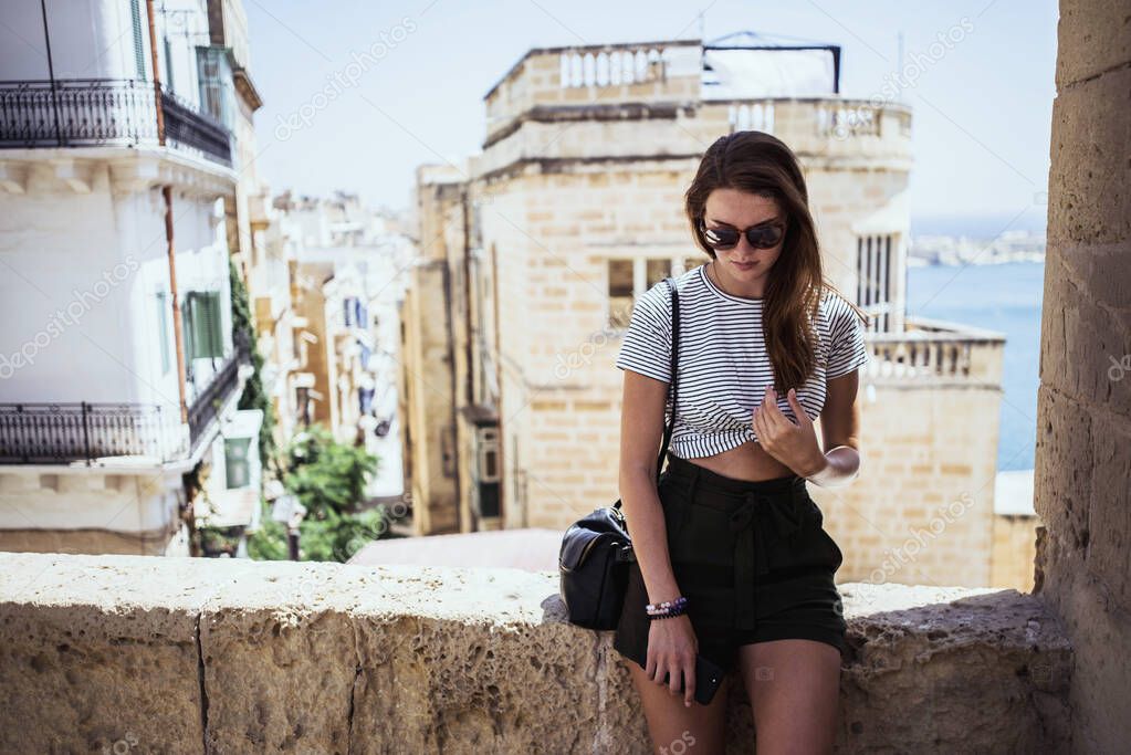 Woman with glasses sitting on the wall. In the background is the sea and the old town of Malta