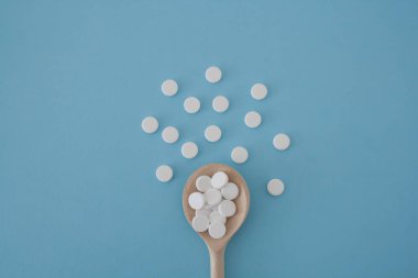 yuvarlak beyaz renkli haplar ahşap bir kaşıkta mavi arka planda, haplar merkezden merkeze dağılmış.