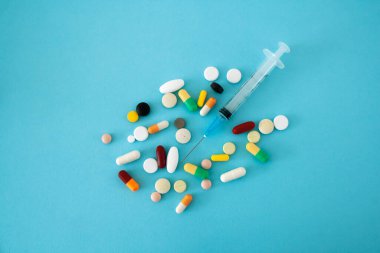 Kavramsal görüntünün merkezinde şırınga bulunan mavi bir arka plana yerleştirilmiş çok renkli haplar ve kapsüller.