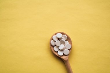 Sarı arka planda tahta kaşıkta kırmızı bir kapsül bulunan yuvarlak beyaz tabletler, kaşık en alttan en üste yerleştirilmiştir.