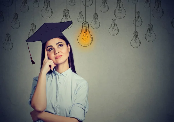 Graduado estudante mulher em vestido de boné olhando para cima em brilhante lâmpada pensando — Fotografia de Stock