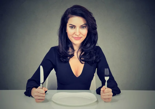 Happy woman with fork and knife sitting at table with empty plate — Stock Photo, Image