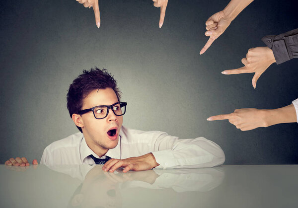 Испуганный работник прячется под столом и его обвиняют люди, указывающие на него пальцем.
 