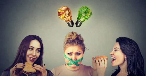 Twee gelukkige vrouwen eten hamburgers kijken benadrukte mollig meisje met meetlint rond haar mond en junkfood en groenten lampen boven hoofd. — Stockfoto