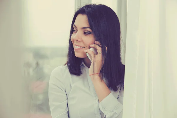 Mujer hablando por teléfono móvil mientras está de pie junto a la ventana en el apartamento —  Fotos de Stock