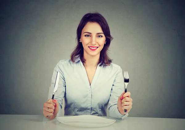 Femme heureuse avec fourchette et couteau assis à table avec plaque vide — Photo