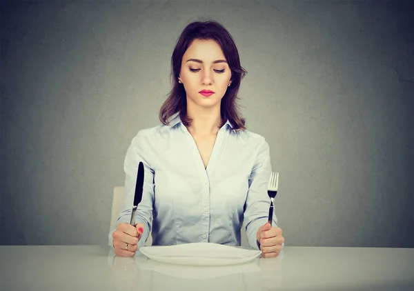 Серьезная женщина с вилкой и ножом сидит за столом с пустой тарелкой — стоковое фото