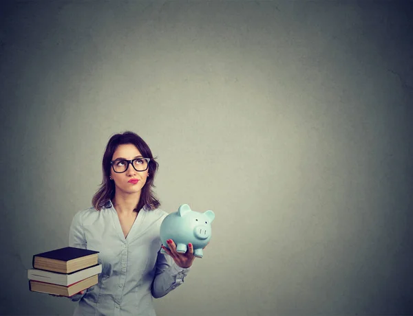 Concepto de préstamo estudiantil. Mujer con un montón de libros y alcancía llena de deudas repensando la futura trayectoria profesional — Foto de Stock