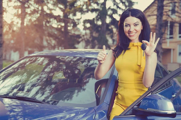 Mulher segurando chaves para carro novo sorrindo mostrando polegares para cima em um fundo de uma casa — Fotografia de Stock