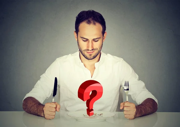 Mann mit Gabel und Messer sitzt am Tisch und blickt auf Teller mit großer roter Frage — Stockfoto