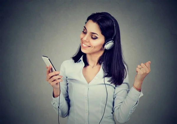 Dançando jovem mulher ouvindo música em fones de ouvido e segurando telefone celular no fundo cinza escuro — Fotografia de Stock