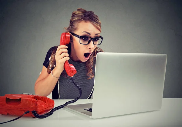 Geschokt jonge vrouw op de laptopcomputer gebruikt op haar Bureau telefoon — Stockfoto