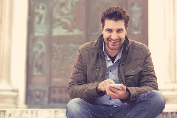 Guapo joven que se relaja en el casco antiguo sosteniendo el teléfono inteligente mirando a la cámara — Foto de Stock