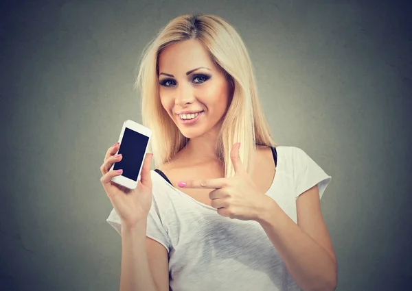 Улыбающаяся девушка указывает на смартфон, стоящий на сером фоне . — стоковое фото