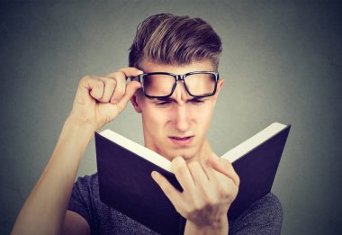 Young man with glasses suffering from eyestrain reading a book having vision problems  clipart