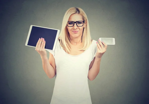 Perplexed young woman in glasses shrugging shoulders uncertain what to choose tablet or smartphone — Stock Photo, Image