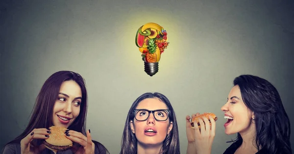 Duas mulheres jovens comendo hambúrgueres olhando para a menina pensativo estressado com frutas em forma de lâmpada acima da cabeça . — Fotografia de Stock