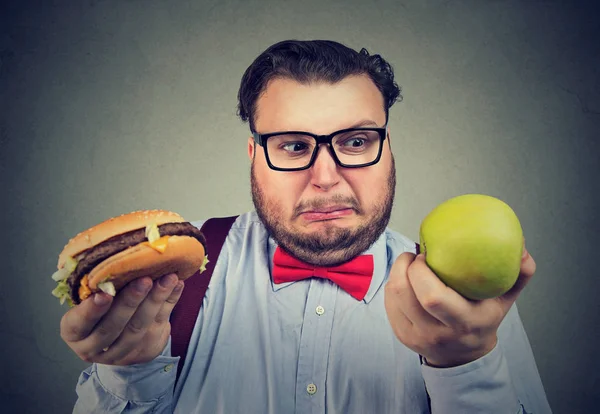 Hombre con manzana y hamburguesa buscando confundido —  Fotos de Stock