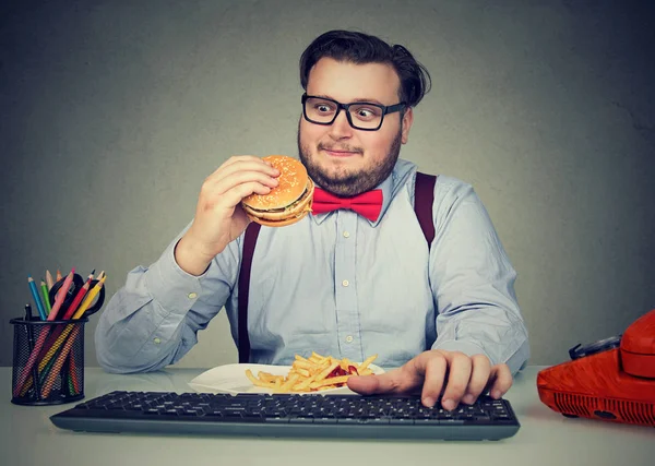 Esfomeado homem robusto comer hambúrguer no local de trabalho — Fotografia de Stock