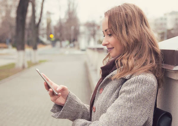Bonita mujer en abrigo sentado en la calle usando el teléfono inteligente y sonriendo —  Fotos de Stock
