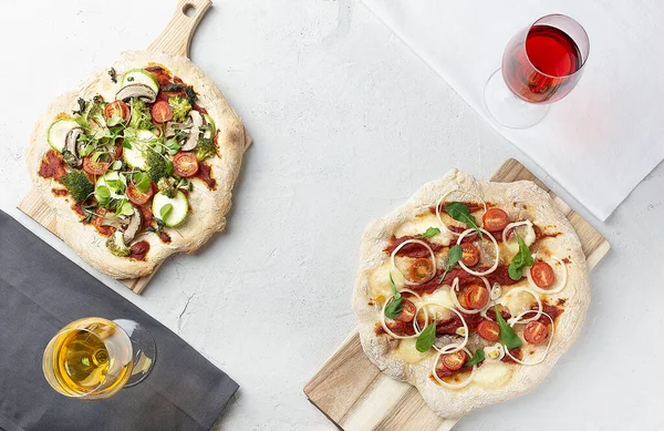 Two types of vegetarian pizza with vegetables on Roman pastry with broccoli, zucchini, tomatoes, mushrooms, microgreens, onions, arugula, mozzarella. On a table. With red and white wine. Top view.