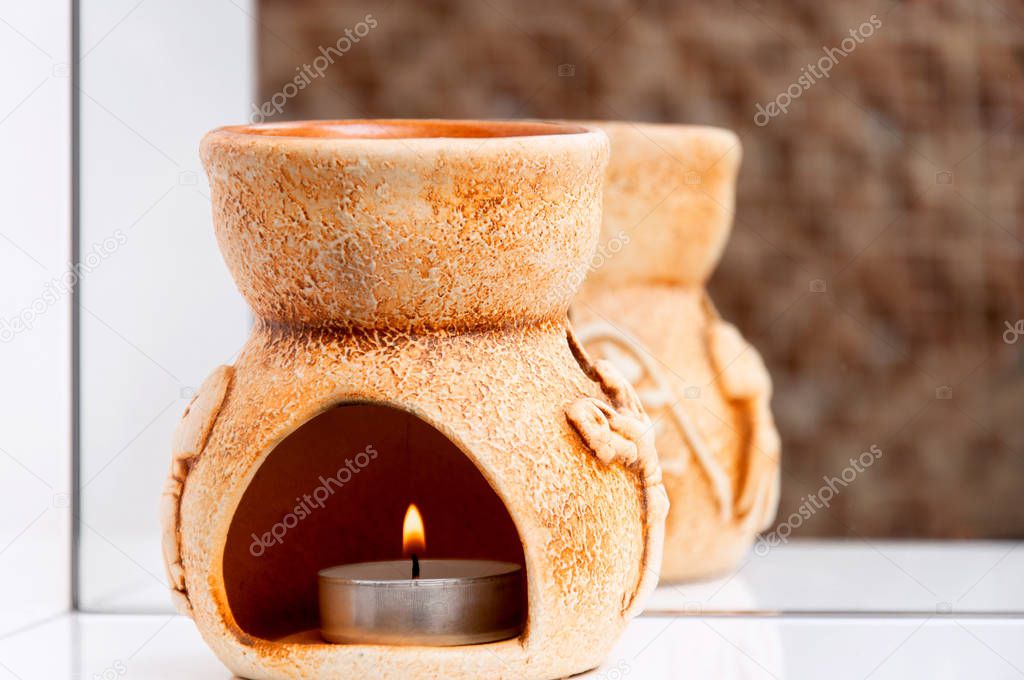 Brown aroma lamp with a burning candle on a white shelf in the bathroom. Personal care for face and body skin. Mirror and shelf in the bathroom with jigsaw puzzle. Relaxation and comfort in the house.