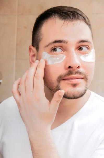 A young guy looks at himself in a mirror with silver patches under his eyes. Handsome guy in the bathroom makes himself a mask. Personal care. Morning routine.