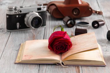 Gri ahşap bir masanın üzerinde, kameranın arka planında duran güzel bir kırmızı gül, eski bir kamera ve açık bir kitap var. Eski kamera, çanta ve filmler gri tahta bir masada.. 