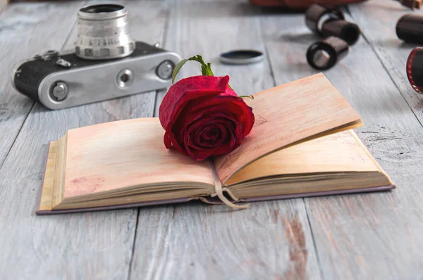 A beautiful red rose lies on a gray wooden table against the background of films from the camera and an old camera and an opened book. Old camera, bag and films on a gray wooden table.
