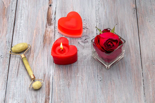 Massage, jade roller for the face, a burning candle in the shape of a heart, a rose in a glass cup on a gray wooden table. Valentine's Day. Personal care. Relaxation, aromatherapy at home.