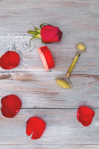 Massage, jade roller for the face, a burning candle in the shape of a heart, a rose in a glass cup on a gray wooden table. Valentine's Day. Personal care. Relaxation, aromatherapy at home.