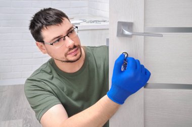 Adam yeni kapı kolları takıyor ve odanın kapısını kilitliyor. Evde tamirat var. Mavi eldivenli adam kapı kollarını tutuyor. Kapı kilidi takılıyor.