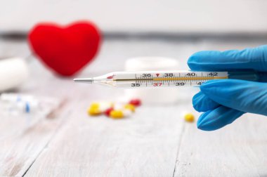 Eldivenli bir kız elinde termometre tutuyor. Termometre yakın. Sargı bezi, haplar, şırınga ve kalp gri ahşap arka planda. Sağlık hizmetleri. Ambulans çağırın. Sağlık ve İlaç Hizmetleri.