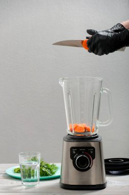 Kız smoothie için havuç kesiyor. Blender, havuç, maydanoz, gri ahşap bir masada dereotu. Kız sebze püresi yapıyor. Sağlıklı bir yaşam tarzı. Beslenme düzeni. Havuç ve yeşillik yakın plan..
