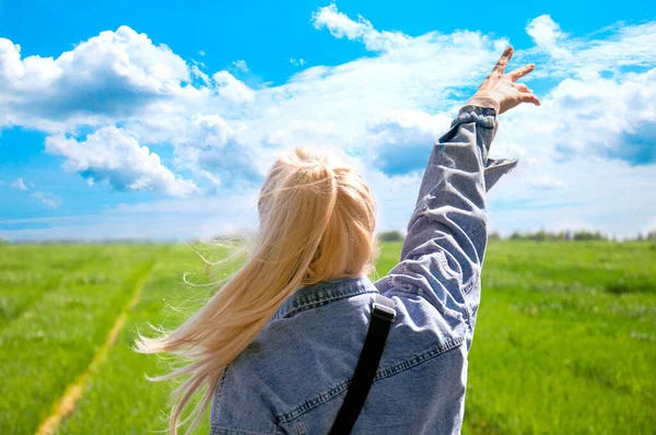 Genç bir kız tarlaya bakıyor. Güzel mavi gökyüzü. Arkaplan yeşili ve gökyüzü. Sarışın, eli kot ceketli bir şekilde gökyüzüne uzanıyor. Güzel kız doğaya hayran..